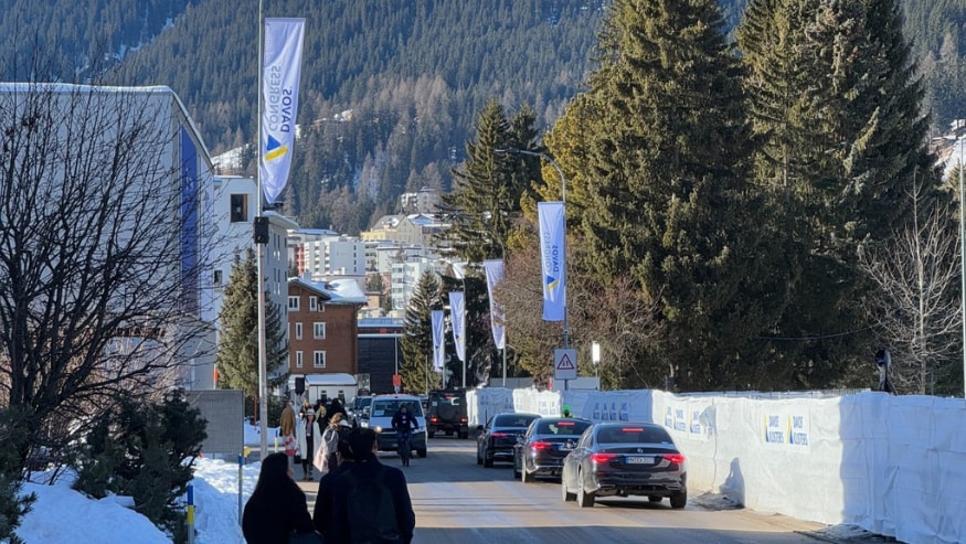 A street scene in a snowy mountain town with several cars and pedestrians. Flags with 'Davos Congress' branding are displayed along the street."