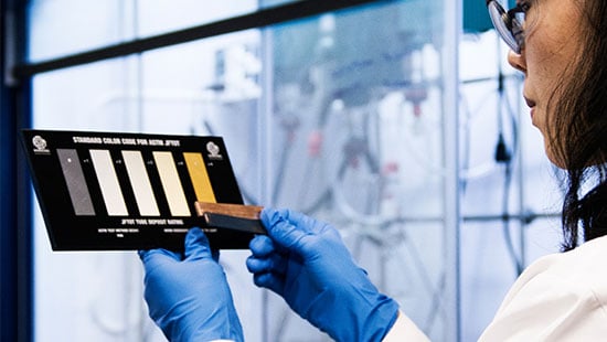 Laboratory chemist wearing protective equipment handling copper pieces against a corrosion reading chart.
